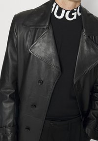Schwarzer Leder-Trenchcoat mit breiten Revers und doppelt geknöpften Knöpfen, getragen über einem schwarzen hochgeschlossenen Oberteil mit weißem Branding.