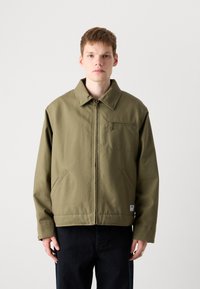 Veste vert olive avec fermeture éclair à l'avant, col pointu, poche poitrine et poches latérales. Fabriquée en tissu durable avec une coupe décontractée.