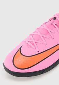 Baskets de sport synthétiques roses avec une surface texturée, présentant des accents noirs et un logo Nike swoosh orange sur le côté.
