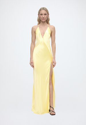 Žena v bezrukávových žltých saténových šatách s hlbokým výstrihom do V, s vysokým rozparkom na stehno a čiernymi sandálmi na remienky, stojaca pred jednoduchým pozadím.