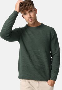 Pull en laine vert foncé tricoté avec des textures horizontales, des poignets côtelés et un col rond. Présente des motifs distincts dans différents styles de tricot.
