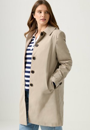 Femme aux cheveux longs portant un manteau beige boutonné par-dessus une chemise rayée bleu marine et blanche, avec une main dans la poche du manteau, regardant sur le côté.