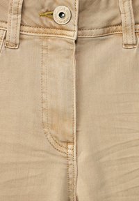 Tejido de mezclilla color beige con un ligero brillo, que presenta un botón de metal con marca grabada, costura central y dos bolsillos laterales.