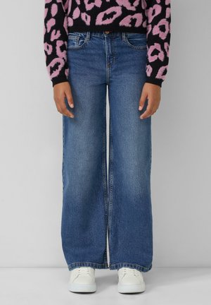 Persona vestida con pantalones vaqueros de pierna ancha azul, zapatillas blancas y un suéter de estampado de leopardo negro y rosa, de pie contra un fondo sencillo.