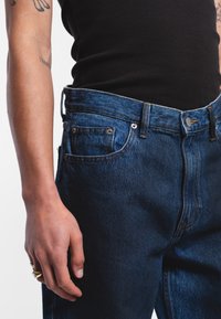 Džínové kalhoty s tmavomodrým omakem, zúženým střihem nohavic a klasickým zapínáním na knoflík. Obsahují přední a zadní kapsy, viditelné stehy.