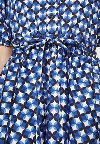 Robe bleu et blanche à motifs avec un design géométrique, dotée d'une fermeture à boutons et d'une taille nouée. Fabriquée en tissu lisse avec une jupe plissée.