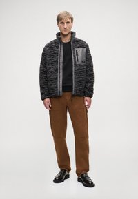 Čierna a šedá textúrovaná bunda na zips s vysokým golierom, bočnými vreckami a šedou časťou. Skombinovaná s hnedými cargo nohavicami a čiernymi topánkami.