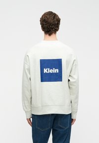 Svetlo šedý sveter s modrým štvorcovým potlačou, na ktorom je v bielom texte v strede na chrbte napísané „Klein“. Mäkko tkaná látka, uvoľnený strih.