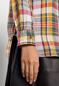Hand resting by side wearing a multicolored plaid long-sleeve shirt and black leather skirt or pants, against a plain background.