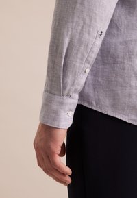 Camicia di lino grigia chiara con polsini abbottonati, che mostra una sottile texture. Presenta un piccolo design ricamato nero sulla manica.