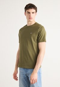 T-shirt olive vert à manches courtes en coton doux, avec un col rond et un petit logo sur la poitrine. Associé à un jean bleu clair.