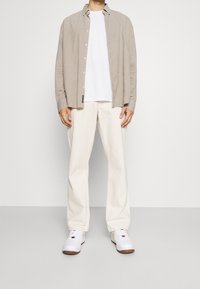 Camisa de manga larga beige claro sobre una camiseta blanca, combinada con pantalones anchos de color crema y zapatillas blancas con suelas marrones.