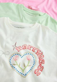 Trzy koszulki z krótkim rękawem: biała z haftem w kształcie kwiatowego serca, zielona o gładkim wzorze i różowa z akcentem w postaci wstążki przy dekolcie.