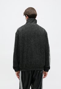 Μαύρο, υφασμάτινο φλις μπουφάν με υψηλό γιακά, που διαθέτει άσπρες πλευρικές ρίγες και χαλαρή εφαρμογή. Χωρίς ορατούς κλεισίματος ή διακοσμητικά στοιχεία.