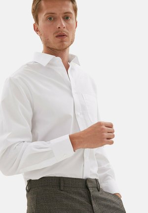 Camicia bianca a maniche lunghe con colletto a punta e taschino sul petto, abbinata a pantaloni grigi a quadri con passante per cintura e vita piatta.