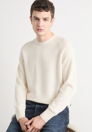 Maglione a maglia color crema con polsini e scollatura a coste. Texture morbida, vestibilità rilassata, indossato con jeans denim scuri. Modella seduta.