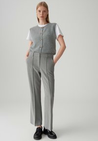 Giovane donna che indossa un gilet grigio con bottoni sopra una camicia bianca a maniche corte, pantaloni grigi a vita alta e scarpe nere con i lacci, in piedi dritta.