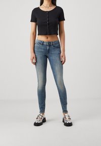 Camiseta negra corta con botones, jeans ajustados azules con acabado desgastado y zapatos de plataforma metálicos con correas ajustables. Acabo con un collar plateado.
