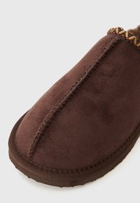 Pantofole in suede marrone con una texture liscia, caratterizzate da cuciture decorative e un motivo accentato lungo il colletto. Design con punta arrotondata.