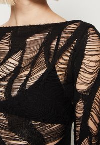 Top nero in maglia con pannelli trasparenti e testurizzati che presentano intricati motivi sovrapposti e un collo a barca. Pelle esposta in alcune sezioni.