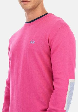 Man in een felroze trui met een klein blauw geborduurd logo op de borst en lichtgrijze patches op de ellebogen tegen een witte achtergrond.