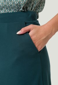 Tyrkysová látková sukně s hladkou texturou. Má boční kapsu, na které spočívá ruka. Na vrchu je vzorovaná blůza v jemných barvách.