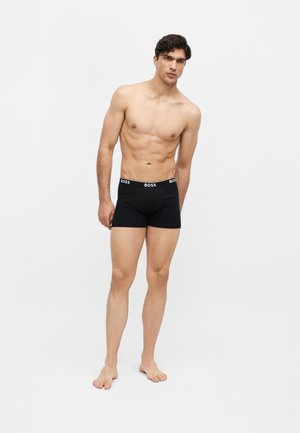Homme torse nu aux cheveux foncés, debout pieds nus, portant un boxer noir BOSS sur un fond blanc uni.