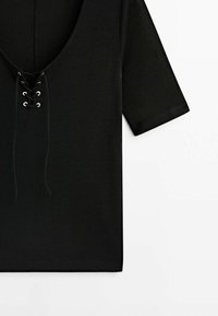 Chemise noire à manches courtes avec laçage sur le col devant et œillets métalliques sur fond blanc.