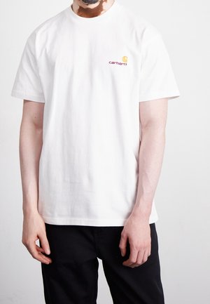 Mężczyzna ubrany w prostą białą koszulkę Carhartt i czarne spodnie, stojący na tle prostego jasnego tła.