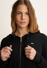 Felpa nera con zip, caratterizzata da una zip dorata, laccetti neri e un distintivo con logo. Il tessuto appare liscio con una leggera lucentezza.
