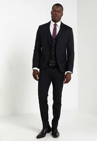 Zwart driedelig pak van gladde stof, bestaande uit een colbert met twee knopen, een vest, slim-fit broek en een patterned das. Gepolijste zwarte schoenen.