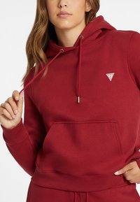 Rode hoodie van zacht materiaal, met een voorzak, geribbelde manchetten en een klein driehoekig logo op de borst.