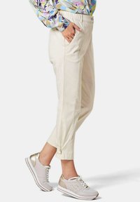 Beige Hose mit entspannter Passform, ausgestattet mit Seitentaschen, einem Knopfdetail am Saum und einem glatten Baumwollmischgewebe. Kombiniert mit hellen Sneakers.