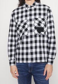 Czarna i biała flanelowa koszula w kratę z dwoma kieszeniami na piersiach, kołnierzykiem na guziki i długimi rękawami. Materiał ma miękką teksturę.