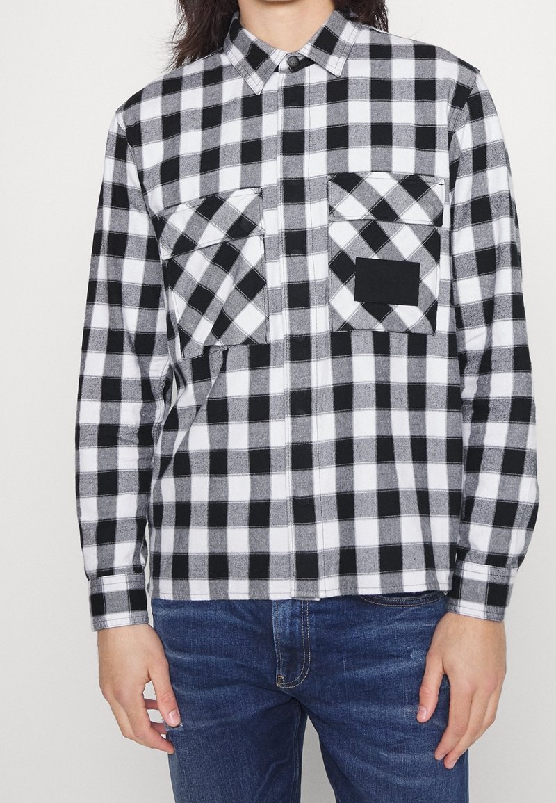 Czarna i biała flanelowa koszula w kratę z dwoma kieszeniami na piersiach, kołnierzykiem na guziki i długimi rękawami. Materiał ma miękką teksturę.