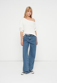 Kremowy sweter z odkrytymi ramionami w plecionkę, zestawiony z jasnoniebieskimi dżinsami o szerokich nogawkach oraz czarno-białymi trampkami. Prosty, swobodny styl.