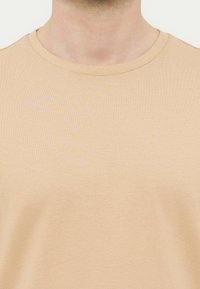 Κοντινό πλάνο ενός ατόμου που φοράει μπεζ μπλούζα με στρογγυλή λαιμόκοψη, εμφανίζοντας τον λαιμό και το πάνω μέρος του στήθους.