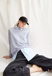 Hellblaues, geschichtetes Hemd unter einem grauen Sweatshirt. Schwarze Kappe mit weißem Logo. Lockere graue Jeans. Weiche Texturen, entspannte Passform.