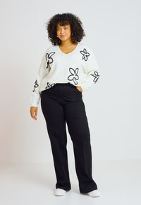 Suéter de punto blanco con patrones florales negros, combinado con pantalones negros y zapatillas blancas sin cordones. El modelo tiene el cabello rizado y se encuentra con una mano en el bolsillo.