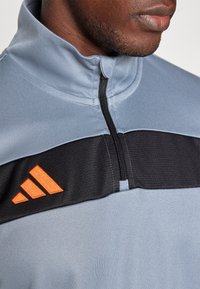 Chaqueta deportiva azul clara con un panel central negro y el logo de Adidas en color naranja, que presenta un alto cuello y cierre de media cremallera.
