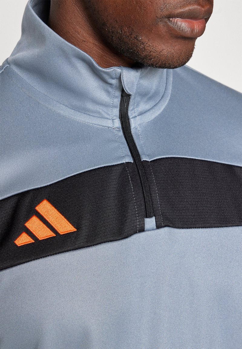 Giacca sportiva azzurro chiaro con pannello centrale nero e logo Adidas arancione, dotata di colletto alto e chiusura a zip parziale.