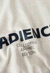 T-shirt in cotone grigio chiaro con grandi scritte in blu navy che recitano "ADIENCE", con un testo più piccolo sottostante che dice "COLLECTIVE APPAREL EDITION."