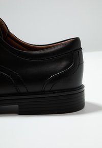 Scarpa in pelle nera con una finitura liscia. Presenta una punta arrotondata e dettagli cuciti lungo i lati. Suola in gomma nera con scanalature.