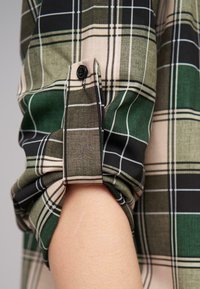 Camicia a scacchi verde e nera con maniche, orlo arrotolato, dotata di un singolo bottone nero e strisce bianche decorative su un tessuto testurizzato.