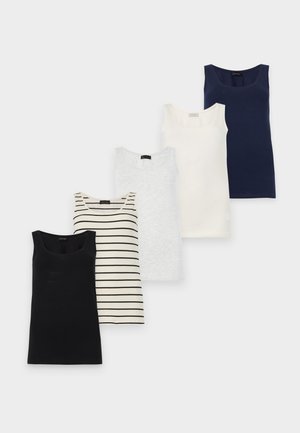 Kvinde står foran en hvid baggrund iført en hvid tanktop, lyseblå bukser med snøre og hvide sneakers, hånd på hoften.