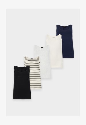 Femme debout sur un fond blanc portant un débardeur blanc, un pantalon bleu clair à cordon et des baskets blanches, la main sur la hanche.