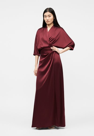 Γυναίκα με μακριά, ίσια μαύρα μαλλιά, ντυμένη με μακρύ κόκκινο σατέν φόρεμα τύπου wrap, με μανίκια τριών τετάρτων και πέδιλα ανοιχτών δακτύλων.