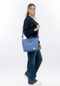 Blauer Tragetasche aus strukturiertem Stoff, mit zwei Seitentaschen und einem Logopatch. Verstellbarer Schulterriemen und ein strukturiertes Design.