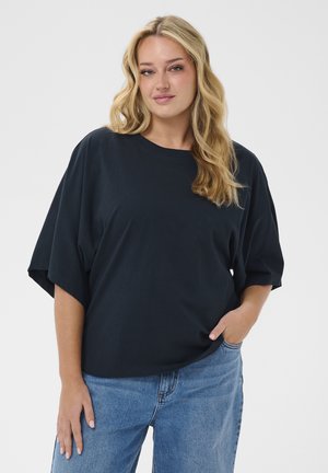 Blondynka o falowanych włosach, ubrana w luźną czarną bluzkę z krótkim rękawem i niebieskie dżinsy, stojąca z jedną ręką w kieszeni na białym tle.