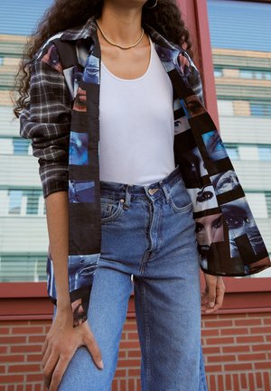 Osoba ubrana w czarną kurtkę z wzorem i kratą, na białym topie na ramiączkach i wysokim stanem niebieskich dżinsów, stojąca przed nowoczesnym budynkiem.